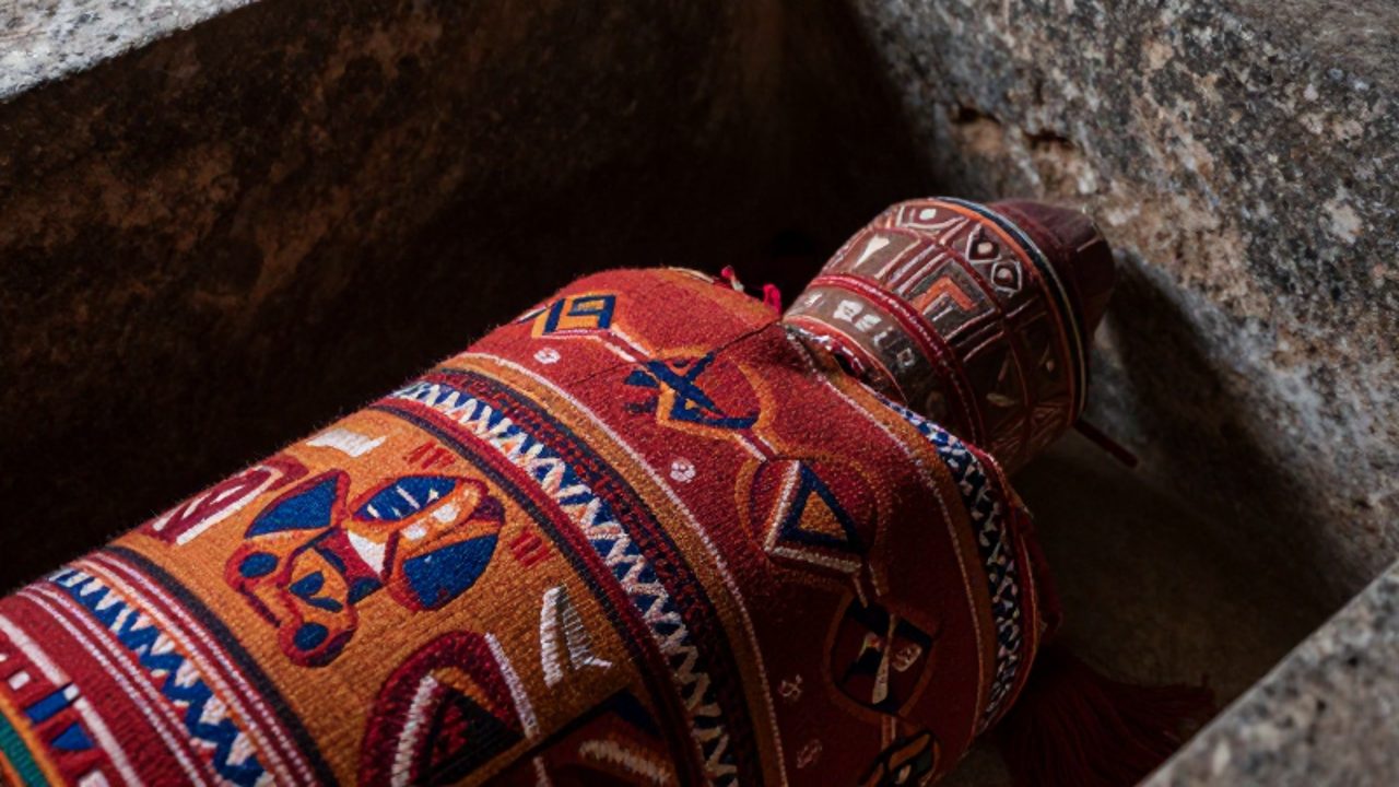 Capas de textiles andinos con patrones geométricos envuelven un recipiente ceremonial dentro de una cámara de piedra en penumbra.